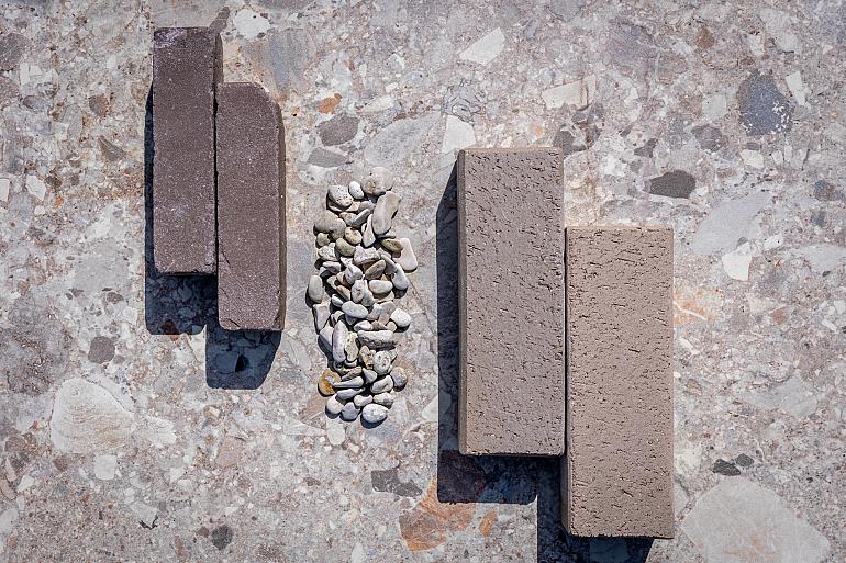 Een bovenaanzicht van vier verschillende bestratingsstenen en een cluster van grind, gepresenteerd op een grotere, gespikkelde terrazzo-stijl tegel. Links liggen twee smallere, donkerder bruin-zwarte stenen (Wienerberger UWF60 Vida - Triton). In het midden bevindt zich een cluster van grijze en witte grindstenen. Rechts liggen twee langere, bredere, beige-grijze gebakken klinkers (Gebakken Klinker XL Colimagrau). De hele compositie is van bovenaf gefotografeerd onder direct zonlicht, met duidelijke schaduwen die naar rechts vallen.